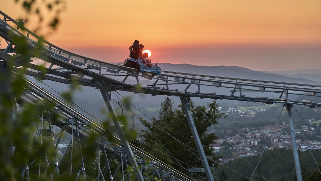 Zwei Personen auf einer Sommerrodelbahn bei Sonnenuntergang mit Bergblick