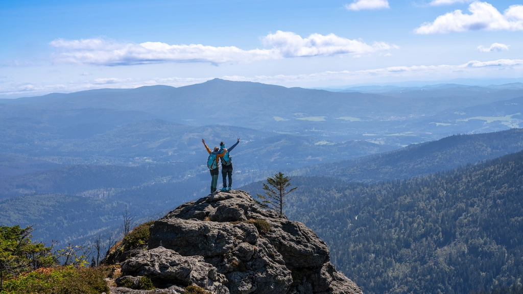 Zwei Wanderer auf Felsen mit Aussicht auf bewaldete Berge und blauen Himmel